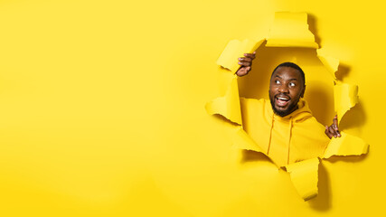 Excited african american man looking at copy space and smiling, showing offer breaking through paper background