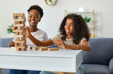 Happy little African American girl with mom taking blocks from tumble tower has fun enjoying communication with parent and playing board game sits on sofa in home environment. Childhood, teenager