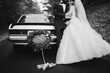 black and white photo of happy young newlywed couple near decorated retro car with plate just married and many jars, outdoors. Bride in white long dress and groom in suit are hugging in Wedding day.