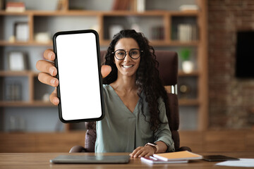 Wall Mural - Positive middle eastern woman sitting at workdesk, showing smartphone, mockup