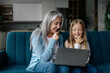 © Prostock-studio - Cheerful caucasian little granddaughter and senior grandma think, watch video on computer, study online