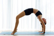 © skumer - Young woman performing wheel yoga pose against a big window with white curtains
