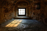Fototapeta Do przedpokoju - Bright sunlight enters the dark stone room through a barred window.
