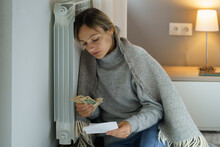 Sad Woman Leaning On Cold Central Heating Radiator Looks At Bills And Holds Money. Lady Covered With Warm Blanket Sits Looking At Debts Without Knowing How To Solve Problem And Prevent Bankruptcy