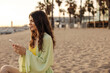 © Look! - The side view of european young woman chatting and looking with smile to phone. Wearing green costume sitting at beach. Use telephone technology concept
