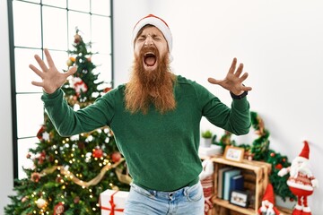 Canvas Print - Redhead man with long beard wearing christmas hat by christmas tree celebrating mad and crazy for success with arms raised and closed eyes screaming excited. winner concept