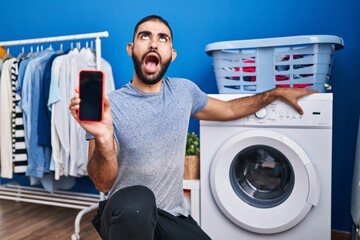 Sticker - Middle east man with beard showing smartphone screen and washing machine angry and mad screaming frustrated and furious, shouting with anger looking up.