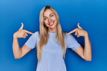 Canvas Print - Beautiful blonde woman wearing casual t shirt over blue background looking confident with smile on face, pointing oneself with fingers proud and happy.