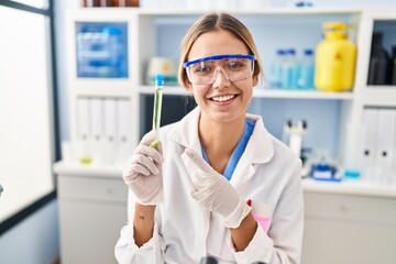 Canvas Print - Young blonde woman working at scientist laboratory holding sample smiling happy pointing with hand and finger