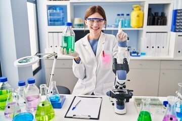 Canvas Print - Young brunette woman working at scientist laboratory smiling with an idea or question pointing finger with happy face, number one