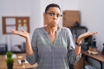 Wall Mural - African american woman working at the office wearing glasses clueless and confused expression with arms and hands raised. doubt concept.