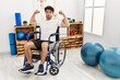 Hispanic man sitting on wheelchair at physiotherapy clinic showing arms muscles smiling proud. fitness concept.