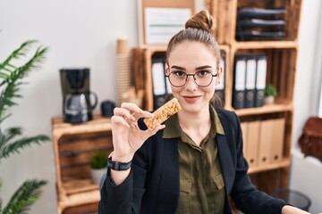 Young hispanic girl eating protein bar as healthy energy snack at the office looking positive and happy standing and smiling with a confident smile showing teeth