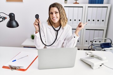 Wall Mural - Young hispanic woman wearing doctor uniform holding stethoscope at medical clinic smiling happy pointing with hand and finger to the side