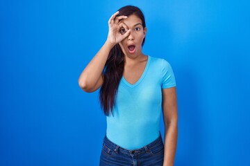 Sticker - Young hispanic woman standing over blue background doing ok gesture shocked with surprised face, eye looking through fingers. unbelieving expression.