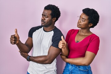 Poster - Young african american couple standing over pink background looking proud, smiling doing thumbs up gesture to the side