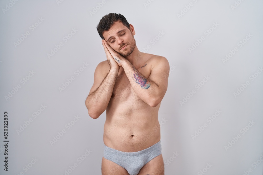 Young hispanic man standing shirtless wearing underware sleeping tired dreaming and posing with hands together while smiling with closed eyes.