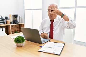 Canvas Print - Senior man working at the office using computer laptop peeking in shock covering face and eyes with hand, looking through fingers with embarrassed expression.