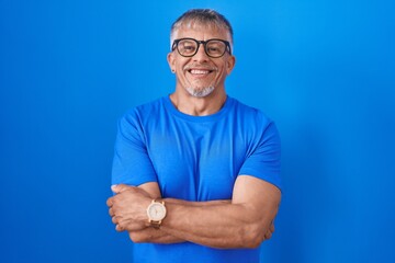 Sticker - Hispanic man with grey hair standing over blue background happy face smiling with crossed arms looking at the camera. positive person.