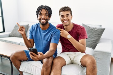 Wall Mural - Young hispanic men using smartphone sitting on the sofa at home with a big smile on face, pointing with hand finger to the side looking at the camera.