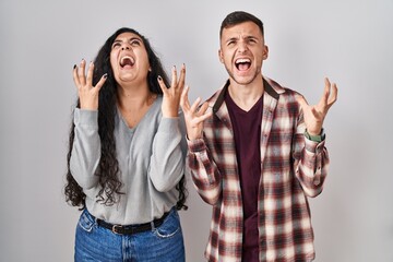 Poster - Young hispanic couple standing over white background crazy and mad shouting and yelling with aggressive expression and arms raised. frustration concept.