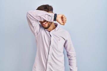 Wall Mural - Arab man with beard standing over blue background covering eyes with arm smiling cheerful and funny. blind concept.