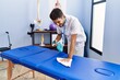 © Krakenimages.com - Young hispanic man wearing physiotherapist uniform disinfecting massage table at rehab clinic