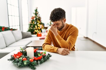 Wall Mural - Arab young man sitting on the table by christmas tree tired rubbing nose and eyes feeling fatigue and headache. stress and frustration concept.