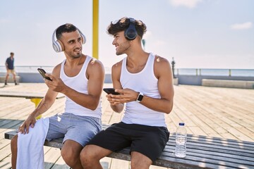 Two hispanic men sporty couple using smartphone and headphones sitting at seaside