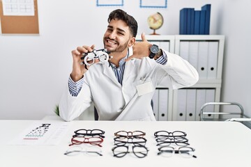 Poster - Young optician man holding optometry glasses smiling doing phone gesture with hand and fingers like talking on the telephone. communicating concepts.