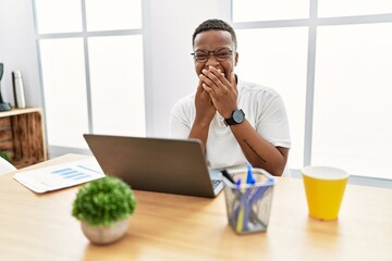 Poster - Young african man working at the office using computer laptop laughing and embarrassed giggle covering mouth with hands, gossip and scandal concept