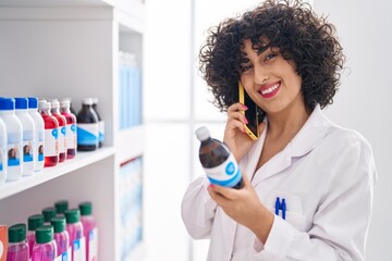 Wall Mural - Young middle east woman pharmacist talking on smartphone holding syrup bottle at pharmacy