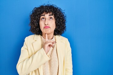 Sticker - Young brunette woman with curly hair standing over blue background thinking concentrated about doubt with finger on chin and looking up wondering