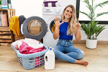 Wall Mural - Young beautiful woman doing laundry sitting by wicker basket showing and pointing up with fingers number six while smiling confident and happy.