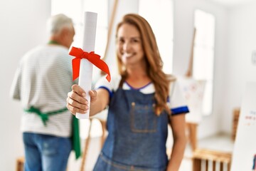 Wall Mural - Two middle age artists smiling happy painting at art studio. Woman standing and holding diploma degree.