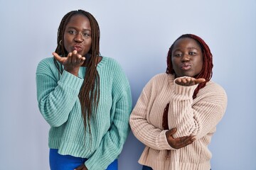 Poster - Two african woman standing over blue background looking at the camera blowing a kiss with hand on air being lovely and sexy. love expression.