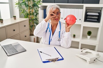 Poster - Middle age woman with grey hair wearing doctor uniform holding balloon smiling happy doing ok sign with hand on eye looking through fingers