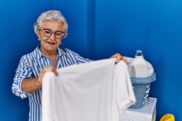 Wall Mural - Senior grey-haired woman smiling confident holding white t shirt at laundry room