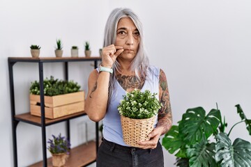 Sticker - Middle age grey-haired woman holding green plant pot at home mouth and lips shut as zip with fingers. secret and silent, taboo talking