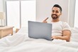 © Krakenimages.com - Young hispanic man with beard using computer laptop on the bed smiling happy pointing with hand and finger