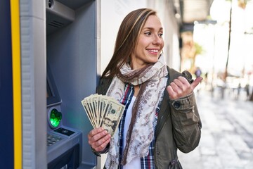 Poster - Young blonde woman holding dollars banknotes from atm machine pointing thumb up to the side smiling happy with open mouth