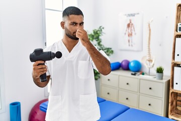 Poster - Young indian physiotherapist holding therapy massage gun at wellness center smelling something stinky and disgusting, intolerable smell, holding breath with fingers on nose. bad smell