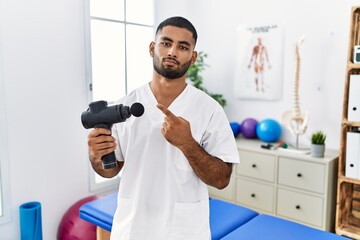 Wall Mural - Young indian physiotherapist holding therapy massage gun at wellness center pointing with hand finger to the side showing advertisement, serious and calm face