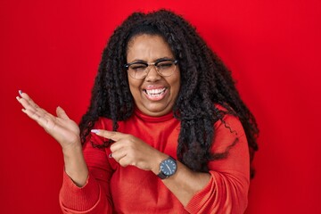 Poster - Plus size hispanic woman standing over red background amazed and smiling to the camera while presenting with hand and pointing with finger.