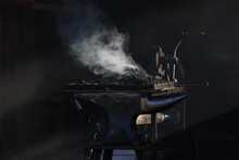 Dark And Moody Image Of A Blacksmith Anvil In Front Of The Forge That Is Beginning To Heat Up As The Pieces Of Coal Catch Fire And Start To Emit Smoke That Catches The Light From An Unseen Window.