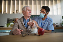 Caregiver With Medical Mask Bringing Healthy Snack To Senior Man In Nursing Home Care Center.