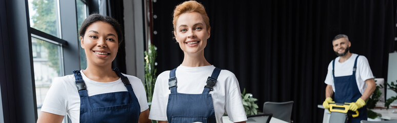 Wall Mural - cheerful interracial women in overalls looking at camera near colleague on blurred background, banner.