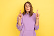Portrait of hopeful woman standing with crossed fingers, wishes her dream comes true, keeps eyes closed, wearing purple hoodie. Indoor studio shot isolated on yellow background.