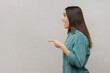 Side view of amazed young adult woman standing, looking ahead with surprised face and pointing at wall copyspace, wearing casual style jacket. Indoor studio shot isolated on gray background.