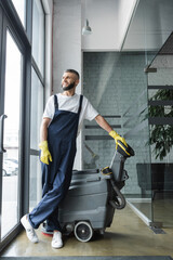 Wall Mural - full length of smiling man in workwear looking away near professional floor scrubber machine.